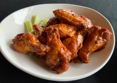 White bowl on a black table. Bowl is filled with chicken wings in an orange/red colored sauce with a few celery stick on the upper right corner of the bowl
