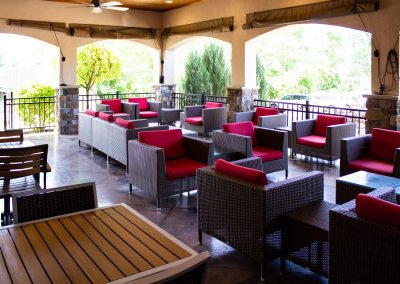 Covered patio that has four open air arches letting in sunlight and air. Outdoor lounging chairs with red cushion line the walls and are spaced through out the patio.