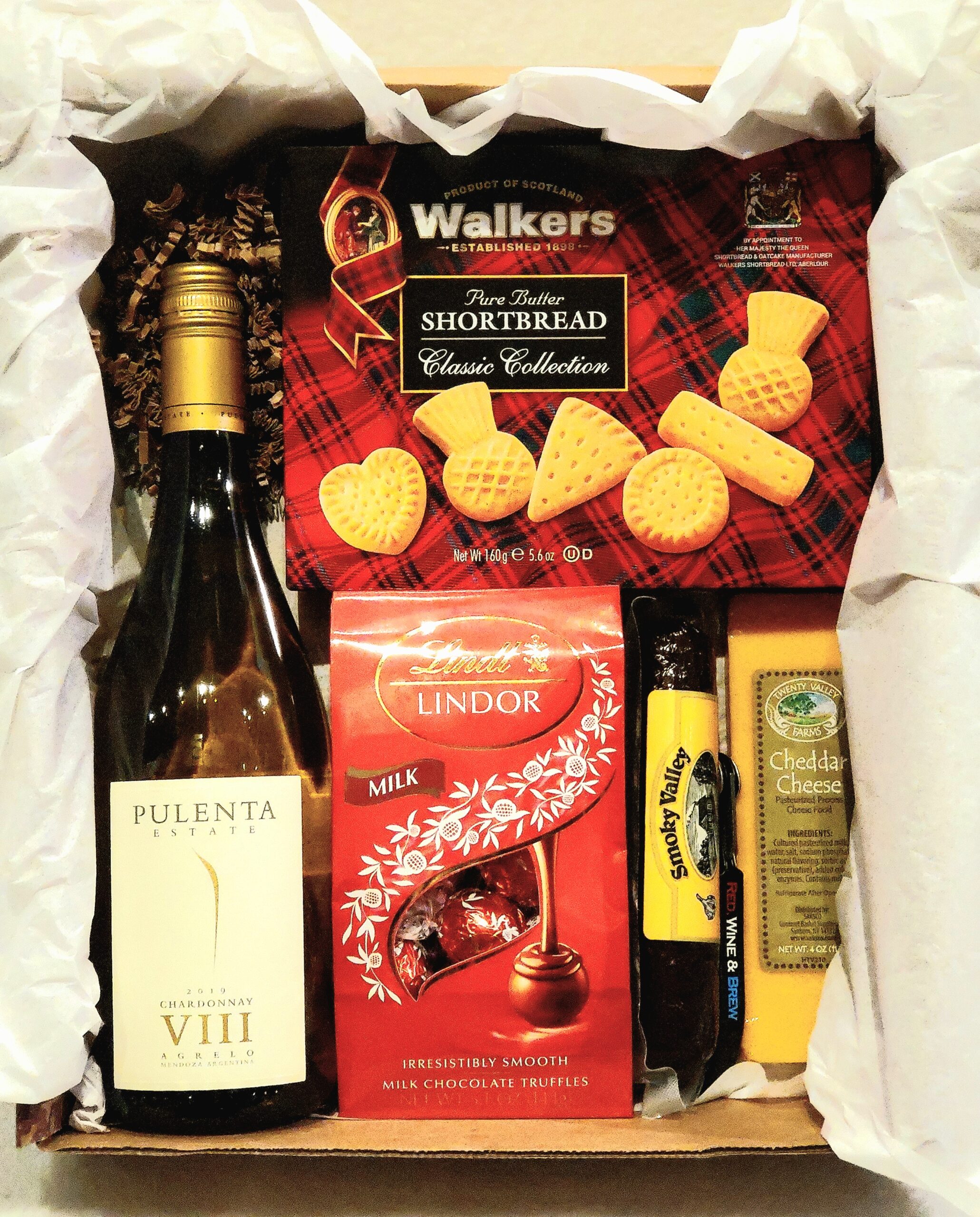 A box filled with crinkle cut paper with a white wine bottle, chocolates, sausage, cheese, and crackers.
