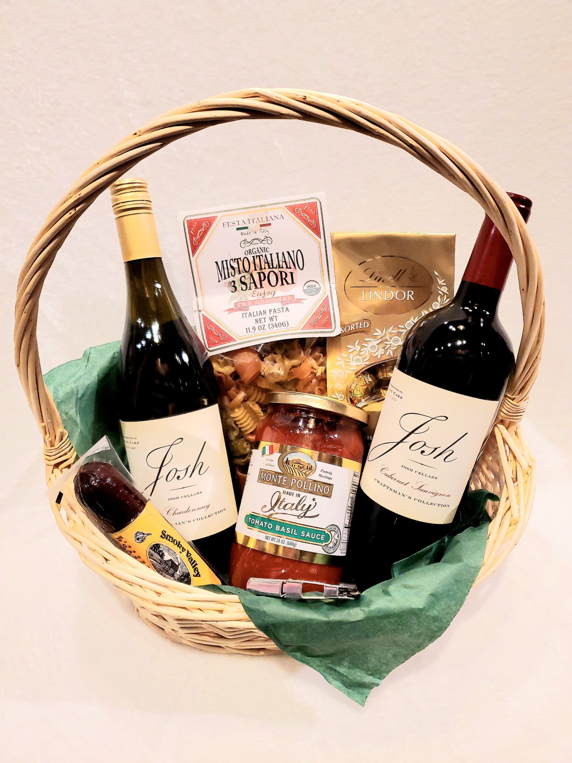 A wicker basket with handle two bottle of wine, pasta, and assorted snacks.