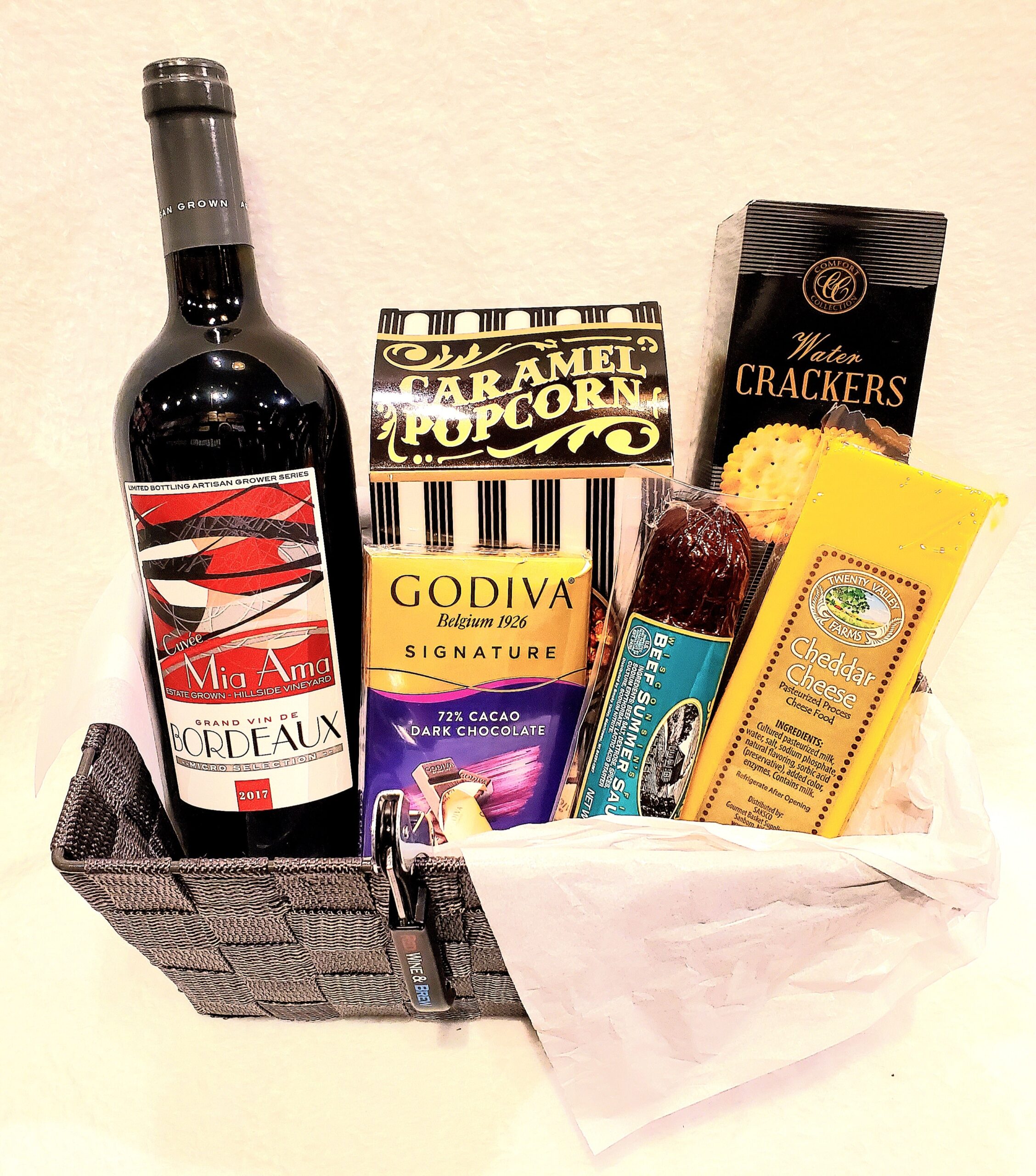 A small woven fabric basket with one bottle of wine and assorted snacks.